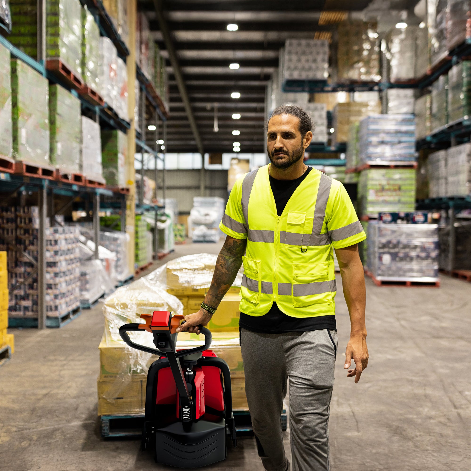 Man in a warehouse wearing a high-visibility vest, pushing a pallet jack.