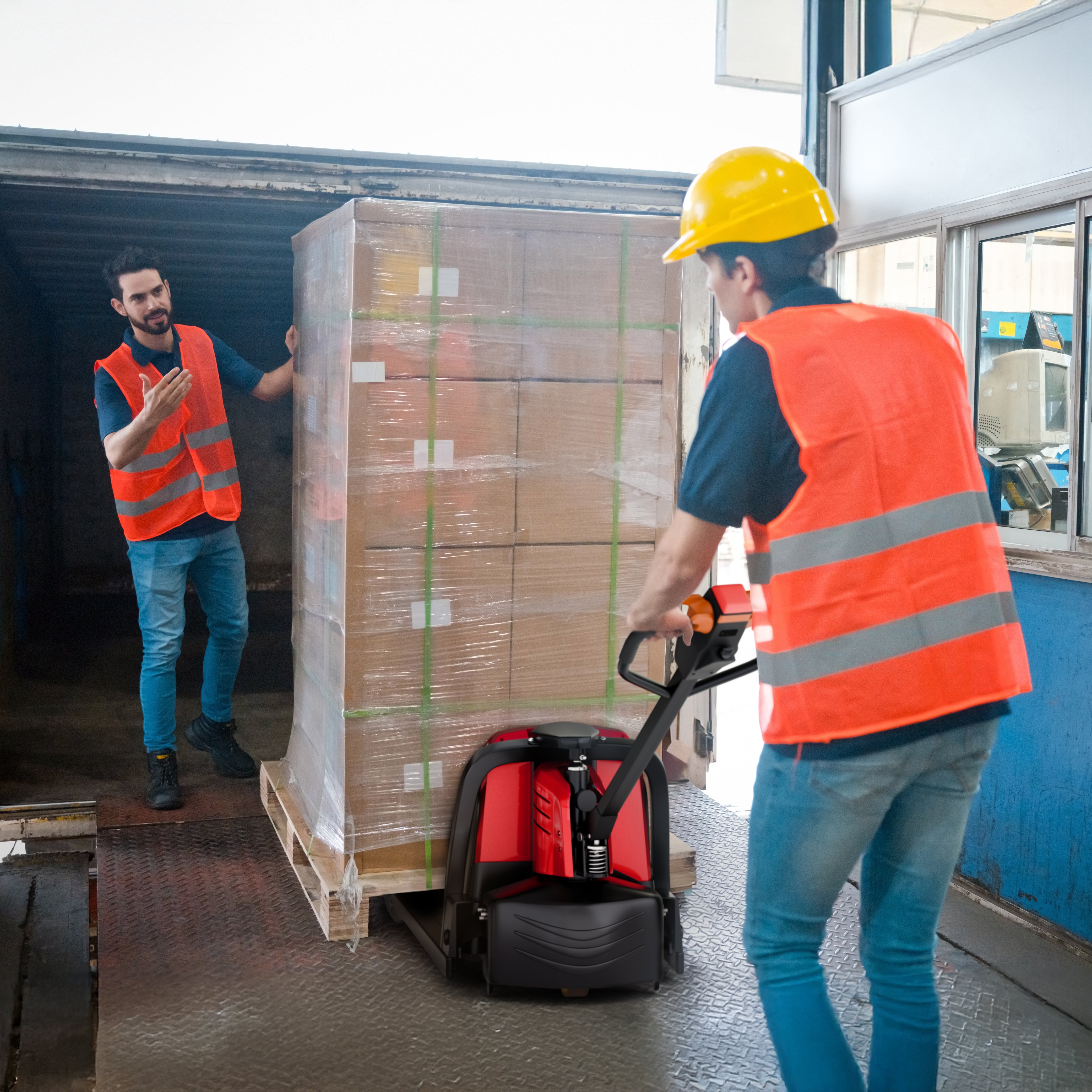 Two workers in high-visibility vests and hard hats moving a pallet of boxes with a hand truck.